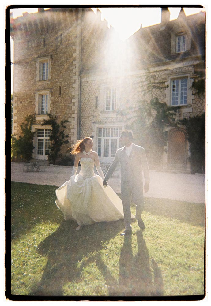 Analog Foto: Brautpaar läuft Hand in Hand über die Rasenfläche vor einem Schloss in Frankreich im Gegenlicht.